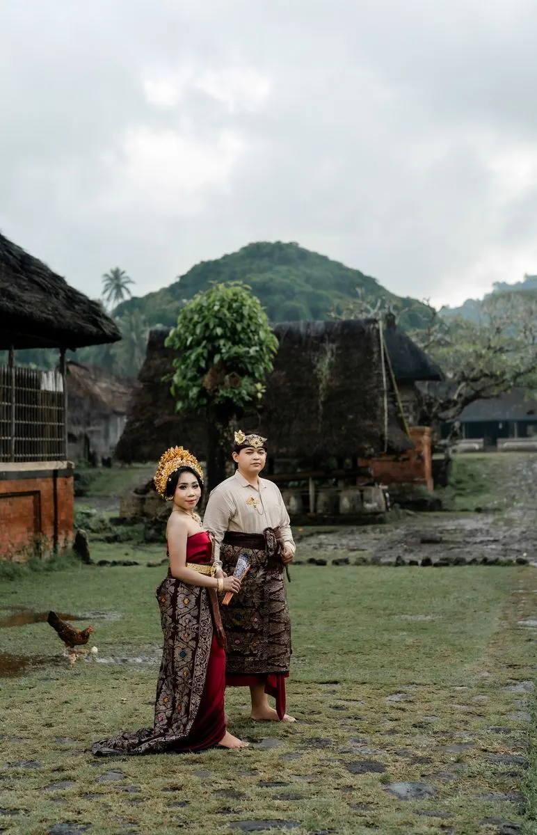 Kedua mempelai berdiri di halaman desa dengan bukit di belakang